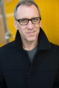 Author Drew Chapman wearing a black overcoat with the collar turned up and standing in front of a mustard yellow bankground