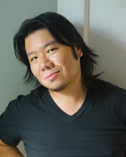 Kevin Kwan wearing a black v-neck t-shirt and with his long black hair loose and flowing around his shoulders