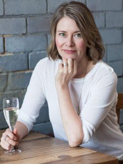 Author Larissa Dubecki wearing a white three-quarter sleeve length top, holding a glass of champagne, sitting at a table in front of a grey brick wall