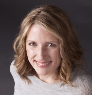 Headshot of author Stephanie Clifford from slightly above. She is wearing a grey jumper and she has blonde shoulder length hair