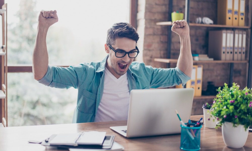 A male author in his office wearing glasses, looking at his laptop and pumping his fists, cheering in celebration.